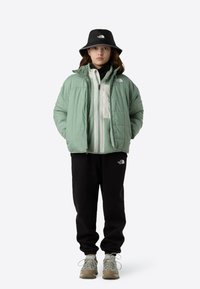 Mintgrøn puffjakke lagdelt over en hvid fleece, sorte joggingbukser og en sort bucket hat. Grå og beige sneakers fuldender outfittet.