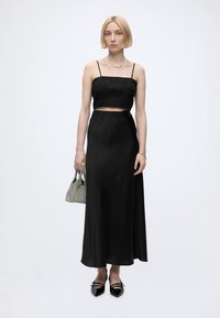 Femme blonde avec coupe au carré porte un haut court en dentelle noire et une longue jupe noire, tenant un petit sac à main texturé et des chaussures noires pointues.