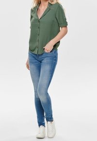 Camicia verde con bottoni, maniche arrotolate, abbinata a jeans skinny azzurri e sneakers bianche. Design semplice e casual con una vestibilità rilassata.