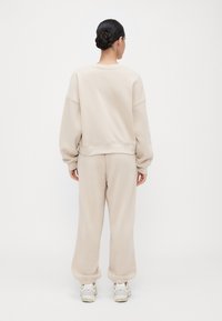 Vrouw met donker haar in een knot, gekleed in een beige oversized sweatshirt en bijpassende sweatpants, staand met haar rug naar de camera tegen een plain witte achtergrond.