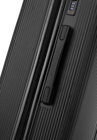 Černý pevný kufr s texturovaným povrchem, horizontálními liniemi a boční rukojetí. Na přední straně má kombinovaný zámek.