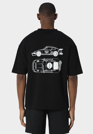 Mand iført sort t-shirt med hvide linjetegninger af en racerbils side- og topvisning på ryggen, stående mod en grå baggrund.