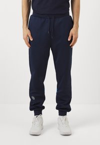 Temno modri trenirki s elastiko v pasu, stranskimi žepki, modrimi črtami in logotipom na spodnjem delu noge. Material iz mešanice bombaža.