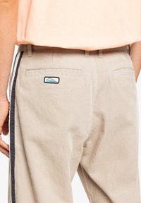 Beige kordfløyelsbukser med en ribbet tekstur, med en svart side stripe og to baklommer. Inkluderer en liten merkelapp over høyre lomme.