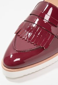 Mocassin en cuir bordeaux brillant avec détail à franges, bout pointu, bordure beige et semelle en caoutchouc blanche sur fond uni.