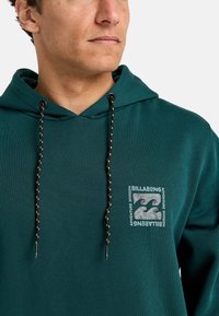 Zielona bluza z kapturem o teksturowanym wykończeniu, czarne sznurki ściągające oraz małe logo graficzne na klatce piersiowej przedstawiające markę Billabong.