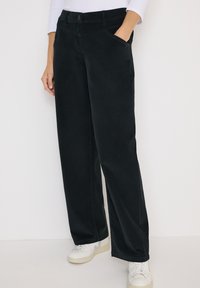 Pantalons larges noirs en tissu doux, dotés de poches latérales, d'une fermeture à bouton et de détails de couture subtils, associés à des baskets blanches.