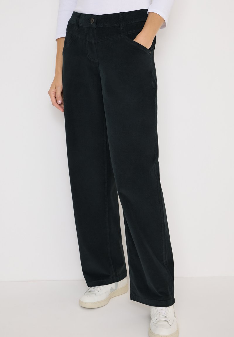 Pantalons larges noirs en tissu doux, dotés de poches latérales, d'une fermeture à bouton et de détails de couture subtils, associés à des baskets blanches.