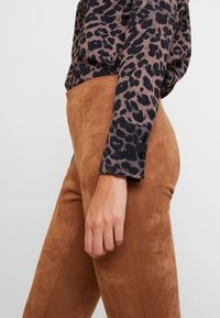 Personne portant un pantalon en suède marron taille haute et une chemise à manches longues imprimée léopard avec des poignets boutonnés, debout devant un fond uni.
