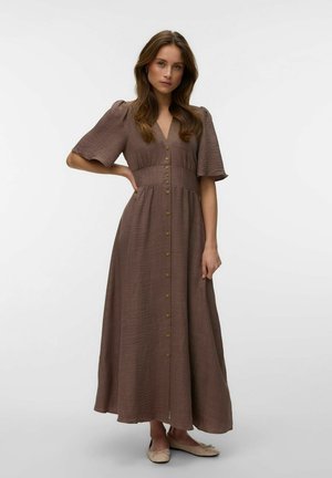 Kobieta stoi z jedną ręką na biodrze, ubrana w brązową sukienkę midi na guziki z bufkami i beżowe baleriny.