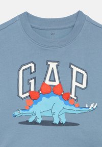 Kék pamut póló egy dinós grafikával, piros és kék tüskékkel, felette vastag fehér betűkkel a "GAP" felirattal.