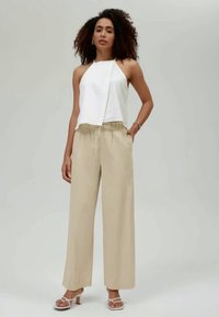 Γυναίκα με σγουρά μαλλιά, φορώντας ένα άσπρο αμάνικο τοπ, μπεζ παντελόνι φαρδιάς γραμμής και άσπρες σαμπάνες με τακούνι, στέκεται μπροστά σε μονόχρωμο φόντο.