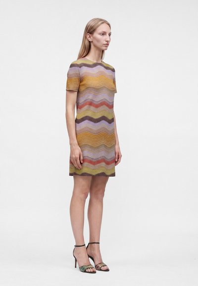 Jeune femme debout portant une robe à manches courtes et à longueur genou avec des rayures horizontales en zigzag multicolores et des sandales noires à talons hauts.