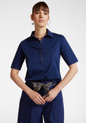 Polo classico blu navy con maniche corte, apertura con bottoni e colletto. Abbinato a pantaloni larghi con una cintura a righe.
