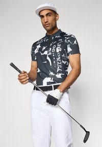 Fekete-fehér grafikai golf póló absztrakt mintákkal, fehér nadrággal és fekete övvel viselve. Golfütőt tart, fehér sapkát és kesztyűt visel.