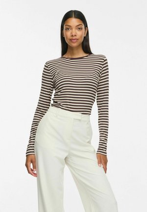 Femme aux longs cheveux noirs portant un haut à manches longues rayé marron et beige et un pantalon blanc taille haute, debout devant un fond blanc.