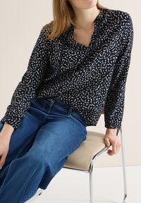 Femme assise sur une chaise beige, portant un haut à manches longues bleu marine à motifs et un jean bleu, les mains reposant sur les cuisses et la chaise.