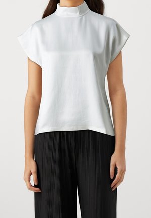 Kobieta w białej bezrękawnikowej bluzce z wysokim kołnierzem i luźnym krojem, w połączeniu z czarnymi plisowanymi szerokimi spodniami, stojąca na tle jednolitego tła.