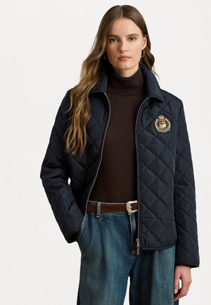 Veste matelassée marine avec fermeture éclair à l'avant, col montant et emblème sur la poitrine gauche. Portée par-dessus un pull col roulé marron et associée à un jean bleu.