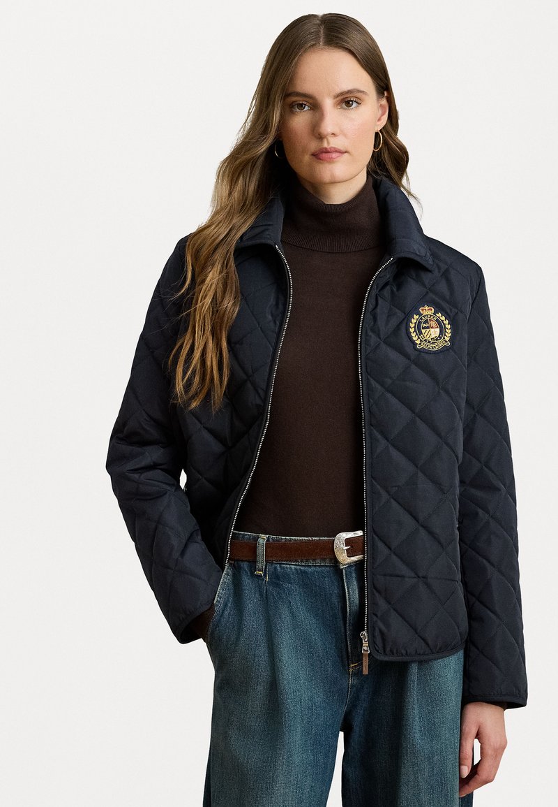 Giacca trapuntata blu navy con zip frontale, colletto alto ed emblema sul lato sinistro del petto. Indossata sopra un dolcevita marrone e abbinata a jeans blu.