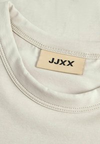 Collo della t-shirt in cotone beige con texture a costine. L'etichetta riporta "JJXX" in nero su uno sfondo crema. Il colletto è rotondo e ben cucito.