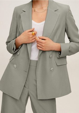 Femme portant un blazer croisé gris clair et un pantalon assorti sur une chemise blanche, avec une bague en or et un collier délicat.