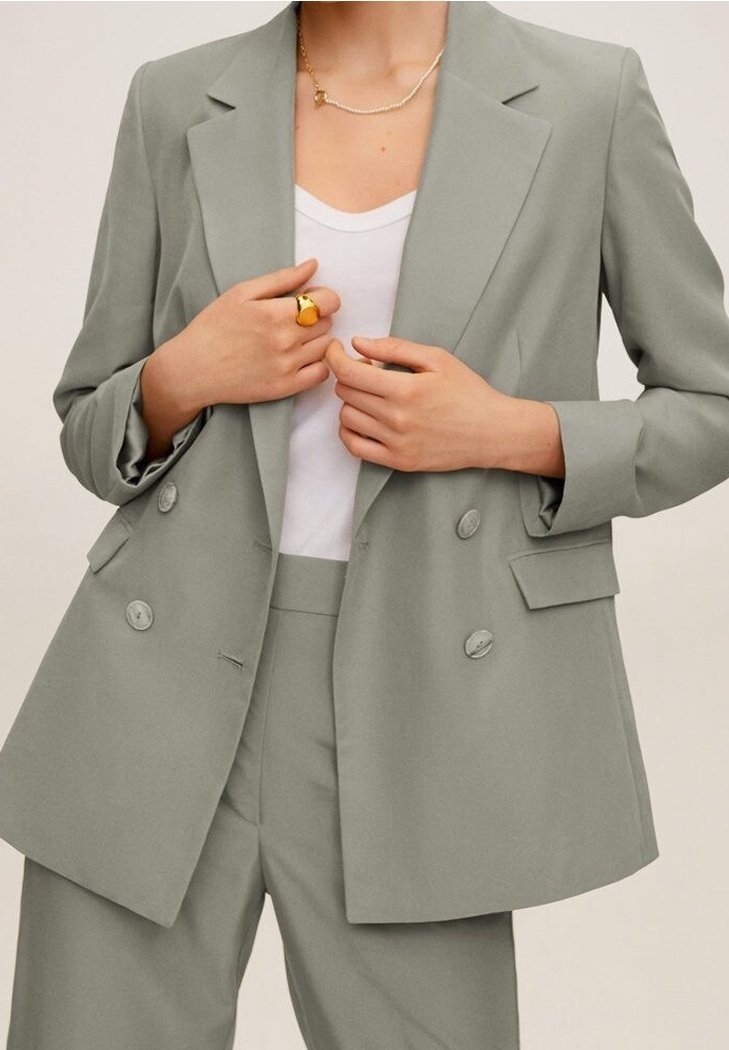 Femme portant un blazer croisé gris clair et un pantalon assorti sur une chemise blanche, avec une bague en or et un collier délicat.