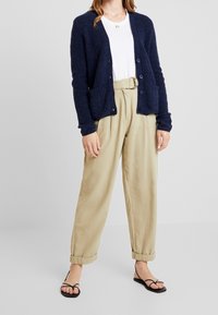 Marinblå stickad kofta med knappar och framfickor, i kombination med beige byxor i hög midja och svarta sandaler med remmar.