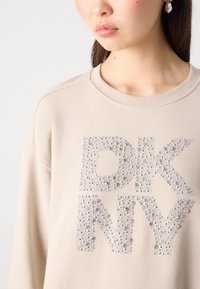 Femme portant un sweat-shirt beige avec de grandes lettres "DKNY" ornées de petits clous argentés sur le devant, visible du cou à la poitrine.