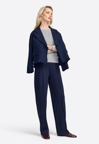 Marineblazer mit strukturiertem Stoff und fransigen Kanten, gestreiftes Langarmshirt und weite Hose. Die Schuhe sind in Dunkelmaron und runden das Ensemble ab.