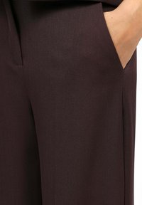 Main droite dans la poche de pantalons sur mesure marron foncé avec une couture visible et une texture de tissu lisse.