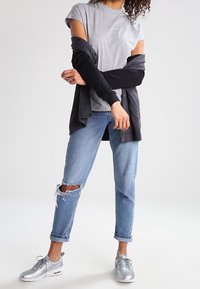 Grå t-shirt, svart cardigan, slitna blå jeans och silverfärgade sneakers. Klädseln har en avslappnad passform med upprullade ärmar och synliga knäsprickor.