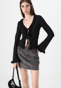 Top negru cu mâneci lungi, cu detalii din volane și închidere cu fundă în față, asortat cu o fustă din denim gri. Modelul poartă o poșetă mică neagră.