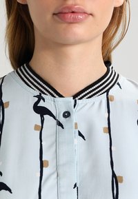 Ljusblå blus med en randig krage i svart och vitt, prydd med ett mönster av svarta fåglar och geometriska former i beige och vitt.