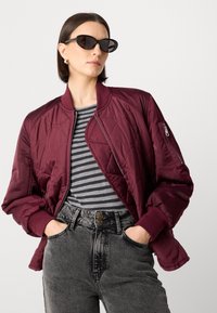 Giacca bomber trapuntata bordeaux con polsini a coste, indossata sopra una maglietta a righe grigie e nere, abbinata a jeans grigi a vita alta e occhiali da sole neri.