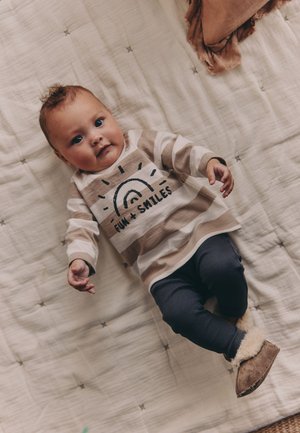 Baby draagt beige gestreept shirt met de tekst "Fun + Smiles", donkere broek en pluizige laarzen, liggend op een lichte gewatteerde deken.