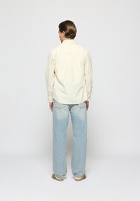 Light yellow button-up shirt with long sleeves, relaxed fit, paired with light blue straight-leg jeans. Smooth fabric and minimal detailing.