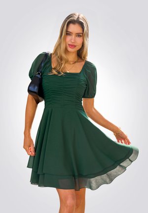 Groene jurk met pofmouwen, een vierkant decolleté, gerimpelde taille en gelaagde rok met een vloeiende silhouet. Gecombineerd met een zwarte handtas.