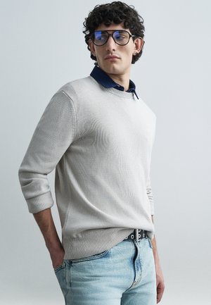 Suéter de punto gris claro con cuello redondo y puños acanalados, usado sobre una camisa azul marino con cuello. Vaqueros azul claro desgastados y cinturón negro.