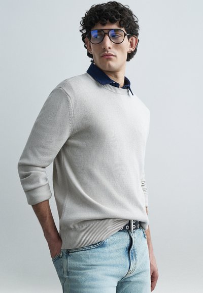 Pull en maille gris clair à col rond, poignets côtelés, porté sur une chemise bleu marine à col. Jean bleu clair déchiré et ceinture noire.