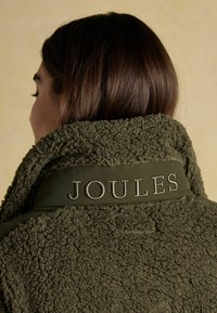 Oliwkowa bluza polarowa o miękkiej fakturze, z wysokim kołnierzem i haftowaną etykietą "JOULES" na plecach.