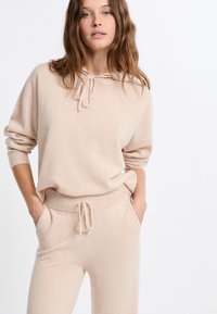 Completo loungewear in maglia beige composto da un pullover con cappuccio e coulisse e pantaloni coordinati con vita elastica e tasche laterali.