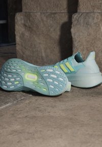 Paar helesiniseid ja kollaseid Adidas jooksujalatseid paigutatud kivipõrandale, näidates talla muster ja külgdisaini.