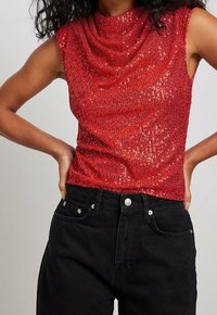 Top sans manches rouge avec un col drapé, fabriqué dans un tissu texturé orné de sequins. Associé à un jean noir taille haute.