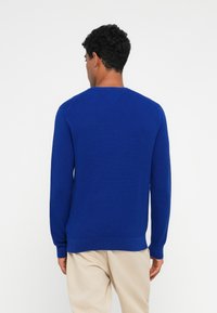 Élénk kék, kötött kerek nyakú pulóver bordázott textúrával és hosszú ujjakkal, tiszta szegéllyel és alakra szabott mandzsettákkal. Puha anyagból készült.