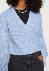 Personne portant un pull en tricot bleu clair par-dessus un haut noir, associé à un jean noir taille haute et un collier court en perles.