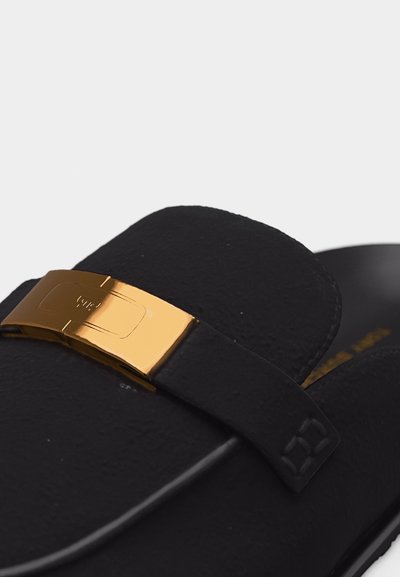 Μαύρο σανδάλι με ευρύ λουρί που διαθέτει γυαλιστερή χρυσή ορθογώνια αγκράφα, εμφανιζόμενο σε κοντινή λήψη με λευκό φόντο.