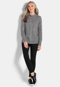 Grauer Strickpullover mit einem Kabeldesign, gerippten Bündchen, getragen über schwarzen Skinny-Jeans und schwarzen Plateausneakern. Einfarbiger weißer Hintergrund.