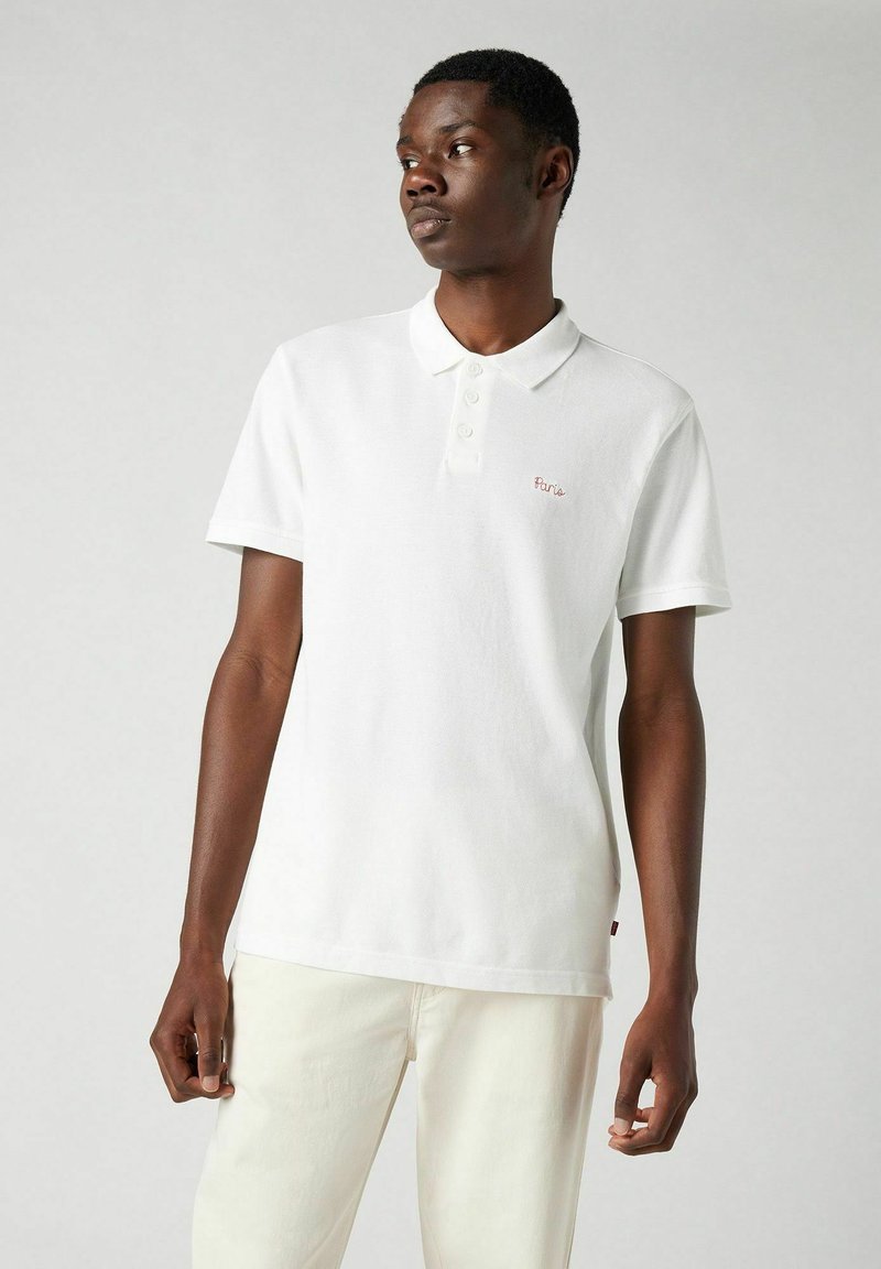 Vit bomullspolo med krage, tre knappar och röd "Paris" brodering på vänstra bröstet. Bärs med off-white byxor.