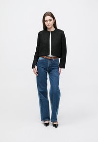 Melnā cropped jaka ar teksturētu audumu, apaļu kakla izgriezumu, bez aizdares, kopā ar zilām izplestām džinsa biksēm un melnām smailām kurpēm.